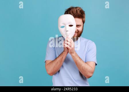 Portrait d'un homme barbu positif qui retire un masque blanc du visage montrant son expression souriante, bonne humeur, prétendant être une autre personne. Studio d'intérieur isolé sur fond bleu. Banque D'Images