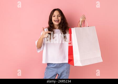 Portrait d'une petite fille excitée portant un T-shirt blanc pointant vers des sacs de shopping, regardant l'appareil photo avec heureux visage expression. Studio d'intérieur isolé sur fond rose. Banque D'Images