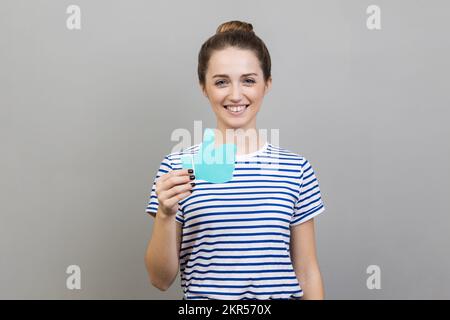 Portrait d'une femme portant un T-shirt rayé montrant les pouces vers le haut du panneau de papier bleu, qui aime et recommandant des postes dans les réseaux sociaux, demandant à évaluer. Prise de vue en studio isolée sur fond gris. Banque D'Images