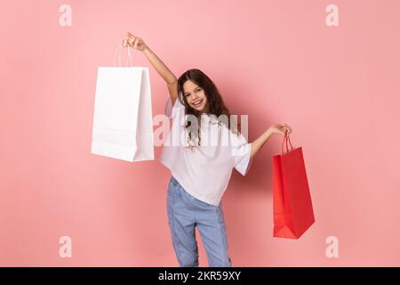 Portrait de la bonne adorable petite fille portant un T-shirt blanc tenant des sacs de papier de shopping, excitée avec des rabais de centre commercial, de bons achats. Studio d'intérieur isolé sur fond rose. Banque D'Images