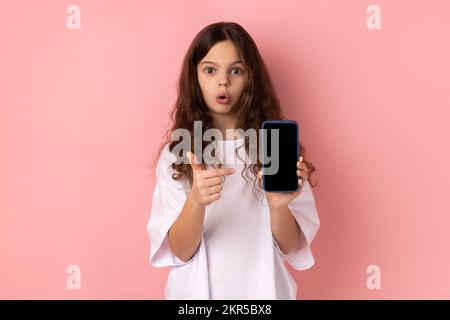 Portrait d'une petite fille surprise choquée portant un T-shirt blanc montrant un smartphone avec écran noir blanc, pointant le doigt sur l'écran. Studio d'intérieur isolé sur fond rose. Banque D'Images