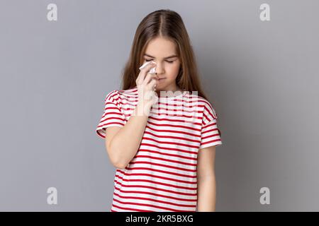 Portrait d'une fillette triste et malheureuse portant un T-shirt rayé se cachant face vers le bas et pleurant, bouleversée par la perte, se sentant chagrin et regret. Prise de vue en studio isolée sur fond gris. Banque D'Images