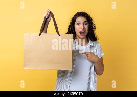 Portrait d'une femme excitée avec des cheveux ondulés foncé tenant et pointant vers le sac de papier, regardant l'appareil photo à la bouche ouverte. Studio d'intérieur isolé sur fond jaune. Banque D'Images