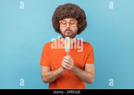 Portrait d'un homme avec une coiffure afro portant un T-shirt orange et des lunettes drôles, tenant la crème glacée, se sent affamé, veut lécher le dessert. Studio d'intérieur isolé sur fond bleu. Banque D'Images