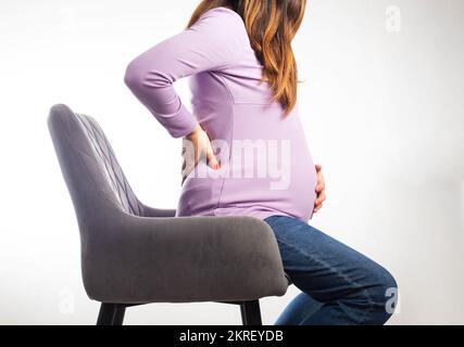 Une fille enceinte s'assoit sur une chaise et la maintient sur un fond blanc. Le concept des maladies de la colonne vertébrale chez les femmes enceintes, les changements hormonaux Banque D'Images
