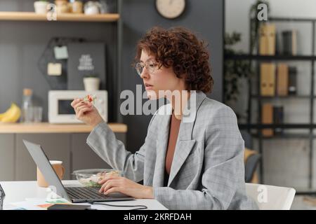 Jeune femme d'affaires assise à table et travaillant sur un ordinateur portable tout en mangeant de la salade pendant l'heure du déjeuner au bureau Banque D'Images