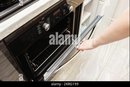 La main d'une femme ouvre la porte d'un four moderne dans la cuisine. Nettoyage catalytique Banque D'Images