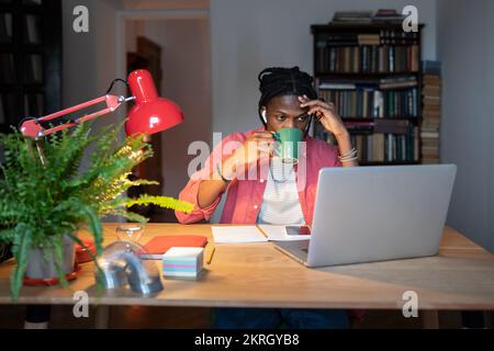 Un étudiant africain fatigué qui étudie en ligne tard dans la nuit à la maison, qui boit du café, se prépare à l'examen Banque D'Images