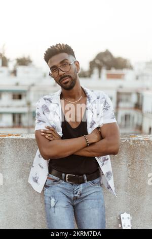 Homme noir en chemise Jean et lunettes avec montre-bracelet sur balcon en béton tout en se tenant avec les bras croisés regardant l'appareil photo sur le plancher élevé de construction Banque D'Images