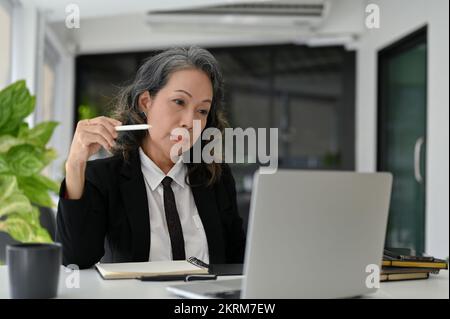 Professionnelle et intelligente femme d'affaires asiatique senior se concentrant sur sa tâche professionnelle sur l'écran d'ordinateur portable, travaillant dans son bureau. Banque D'Images