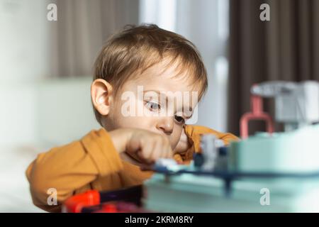 Adorable petit garçon aux cheveux blonds dans un chandail orange assis à table et jouant avec le jouet tout en passant du temps à la maison Banque D'Images