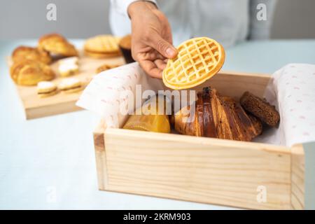 Crop anonyme femme mettant délicieux gaufres maison dans une boîte en bois avec cookie de boisson fraîche et croissant Banque D'Images
