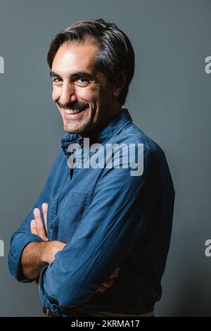 Portrait d'un homme joyeux en chemise bleue souriant et regardant l'appareil photo avec des bras croisés sur fond gris Banque D'Images