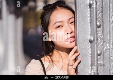 Jeune femme asiatique rêveuse regardant l'appareil photo tout en s'appuyant sur un mur gris au soleil sur un arrière-plan flou de la rue Banque D'Images