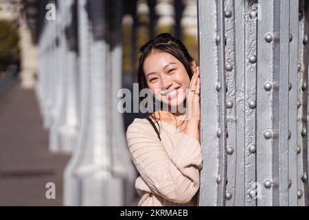 Jeune femme asiatique rêveuse souriante regardant l'appareil photo tout en s'appuyant sur un mur gris au soleil sur un arrière-plan flou de la rue Banque D'Images