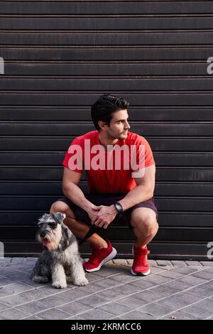 Corps complet d'athlète masculin adulte dans des vêtements de sport rouges assis sur des haunches près de la porte en métal gris avec adorable Schnauzer miniature sur la laisse et regarder loin Banque D'Images