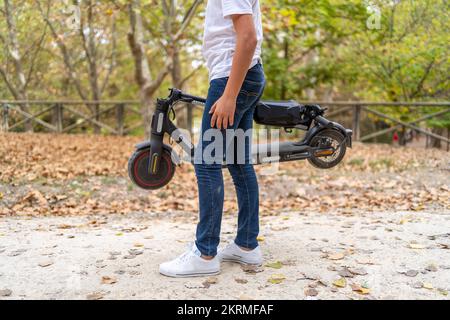 Croquer un adolescent anonyme en t-shirt blanc et jeans debout sur la voie avec scooter électrique dans le parc avec feuillage tombé Banque D'Images