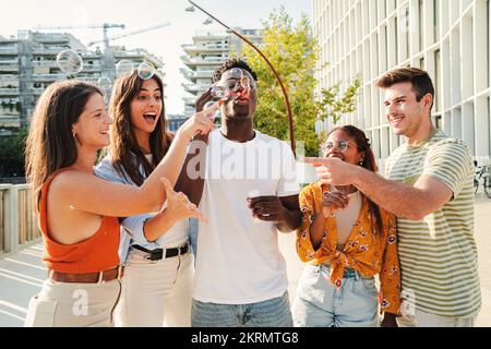 Groupe multiethnique souriant d'adolescents soufflant de la soupe à bulles et appréciant dehors ensemble à côté du campus. Des adolescents gaies et jeunes jouant avec la baguette de bulle le jour ensoleillé dans la rue. Concept de style de vie. Photo de haute qualité Banque D'Images