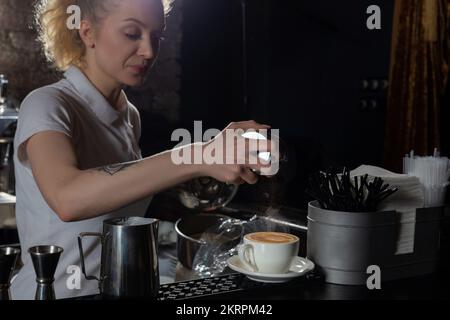 Une femme Barista saupoudrer à la main la cannelle sur le café cappuccino ou le latte sur le comptoir du bar dans le café. À l'aide de la lumière, l'accent est mis sur une tasse de Banque D'Images