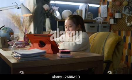 Une jeune fille asiatique s'assoit à la table à la maison confortable et parle avec un professeur. Une écolière écoute la leçon en ligne, discute de nouveaux matériaux et des études sur tablette. Concept d'apprentissage à distance. Conférence à distance. Banque D'Images