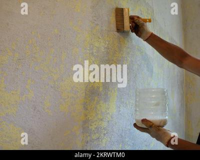 personne tenant une brosse à peinture et un contenant en plastique avec un coulis blanc dans ses mains et appliquant une couche d'apprêt sur un mur épluché texturé Banque D'Images