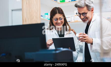 Un pharmacien mature appelle un patient avec une tablette dans un magasin de médicaments. Deux fournisseurs de soins de santé traitant des prescriptions en ligne dans une pharmacie. Banque D'Images