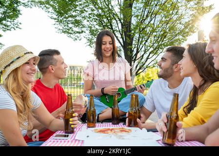 groupe de jeunes étudiants au parc ayant un pique-nique avec pizza et bières, les enfants jouant et chantant avec ukulele et tambourine, réunion heureuse de mille Banque D'Images