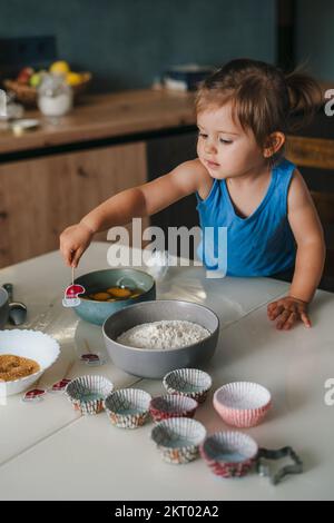 Portrait en gros plan d'une adorable petite fille caucasienne debout devant une table pleine de bols avec des ingrédients préparés pour faire une nouvelle recette de gâteau Banque D'Images