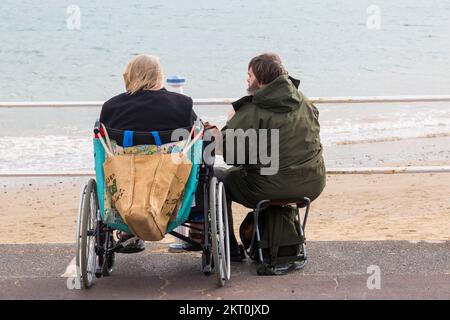 Couple assis sur l'esplanade de la promenade, femme en fauteuil roulant, homme assis sur un siège rabattable, à Weymouth, Dorset, Royaume-Uni, en octobre Banque D'Images
