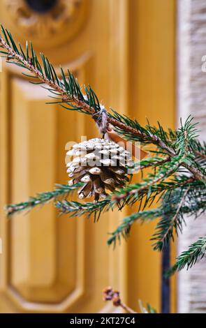 Gros plan de la pomme de pin accrochée à l'arbre de Noël devant la porte jaune vintage. Noël Banque D'Images