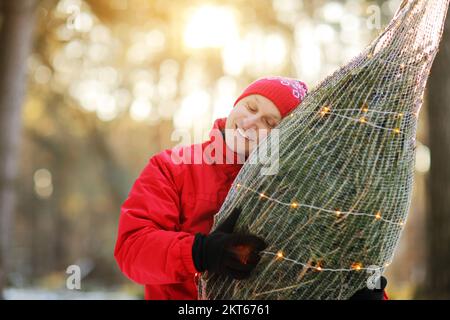 homme souriant portant un arbre de noël fraîchement coupé dans la forêt. Le jeune bûcheron porte un sapin sur son épaule dans les bois. Comportement irresponsable t Banque D'Images