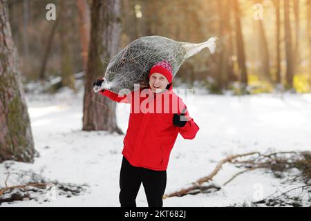 homme souriant portant un arbre de noël fraîchement coupé dans la forêt. Le jeune bûcheron porte un sapin sur son épaule dans les bois. Comportement irresponsable t Banque D'Images