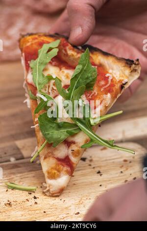Prendre à la main le dernier morceau ou tranche de pizza avec sauce tomate, mozzarella et feuilles de roquette fraîches d'un panneau en bois, gros plan, vertical Banque D'Images
