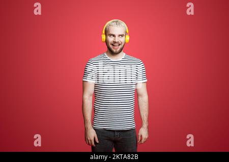 Portrait d'un jeune homme amusant et heureux écoutant de la musique dans un casque, souriant heureux, debout sur fond rouge. Profiter de sa musique préférée. Banque D'Images