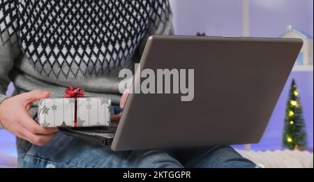 Un homme avec un ordinateur portable et un cadeau achète des cadeaux pour le nouvel an, Noël. Chapeau d'homme à Santa sur fond d'arbre de Noël, guirlandes, cadeaux avec ordinateur portable. Achats en ligne, achats dans une boutique en ligne, Marketplace. Banque D'Images