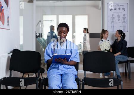 Une infirmière afro-américaine remplit les documents administratifs des patients assis aux chaises de réception de la tour médicale. Un jeune médecin rédige des ordonnances dans la salle d'attente de l'hôpital. Membre du personnel médical qui examine la liste des patients. Banque D'Images