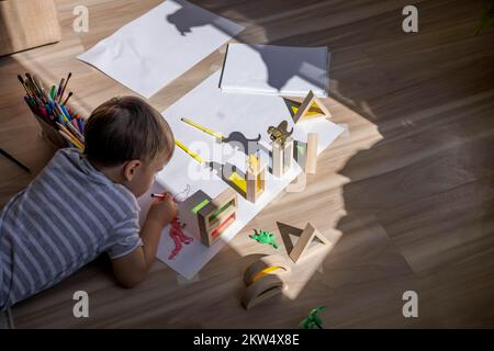 Un petit garçon mignon tire sur le sol de sa chambre. Cercles sur une feuille blanche les ombres des dinosaures projettent de la lumière du soleil. Banque D'Images