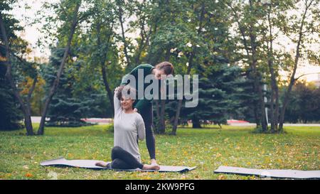 Diligent élève de yoga est d'apprendre la position de visage de vache assis sur le tapis et les bras d'étirement tandis que l'enseignant expérimenté l'aide, les filles parlent et rient à l'extérieur de ptactice. Banque D'Images