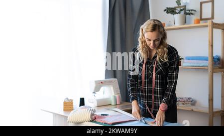 Couturière féminine utilisant du tissu de coupe au couteau sur les lignes d'esquisse dans l'atelier de couture. Des mains de couturier coupant le détail de la robe dans la boutique de tailleur. Copier Banque D'Images