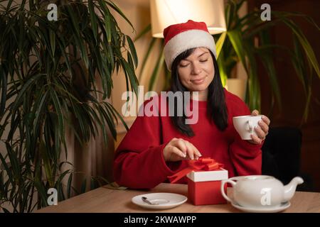 Un brunette dans un chapeau de père Noël boit du thé le matin en déballant un cadeau de Noël. Atmosphère familiale et concept de noël Banque D'Images