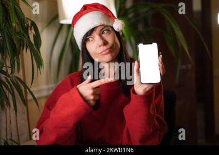 Jeune Santa femme pointant du doigt vers un smartphone mobile écran vide à la veille de Noël. Copier l'espace Banque D'Images