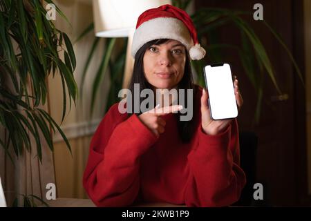 Brunette femme dans le chapeau du Père Noël montrant un écran de smartphone vierge et pointant avec le doigt dessus tout en regardant la caméra. Soldes de Noël en ligne. C Banque D'Images