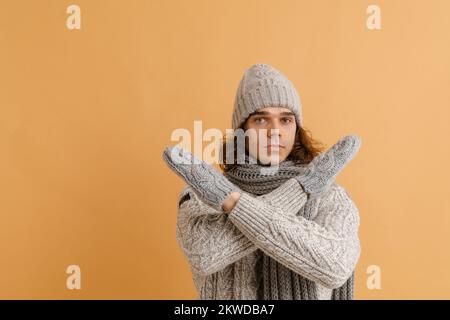 Jeune homme beau avec de longs cheveux en chandail et chapeau d'hiver, écharpe et moufles avec bras croisés sur fond marron isolé Banque D'Images