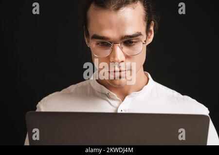 Homme adulte aux cheveux longs dans des lunettes et une chemise tenant un ordinateur portable et regardant sur l'écran sur un arrière-plan noir isolé Banque D'Images