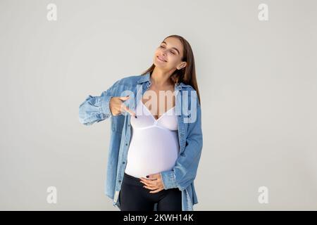 Jeune femme enceinte pointant vers son grand ventre et regardant heureuse Banque D'Images