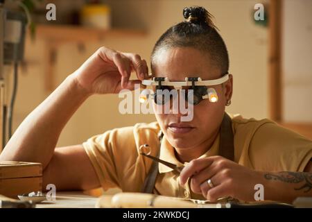 Bijoutier féminin tenant une bague artisanale et portant des lunettes grossissantes dans un studio confortable Banque D'Images