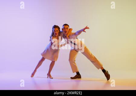 Danse incendiaire. Couple émotionnel de danseurs en tenues rétro dansant social danses isolées sur fond de couleur lilas dégradé dans la lumière de néon Banque D'Images