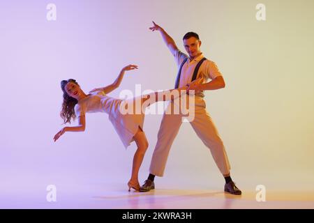 Danse incendiaire. Couple émotionnel de danseurs en tenues rétro dansant social danses isolées sur fond de couleur lilas dégradé dans la lumière de néon Banque D'Images