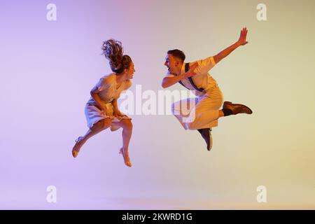 Danse incendiaire. Couple émotionnel de danseurs en tenues rétro dansant social danses isolées sur fond de couleur lilas dégradé dans la lumière de néon Banque D'Images