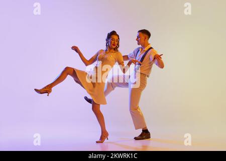 Danse incendiaire. Couple émotionnel de danseurs en tenues rétro dansant social danses isolées sur fond de couleur lilas dégradé dans la lumière de néon Banque D'Images